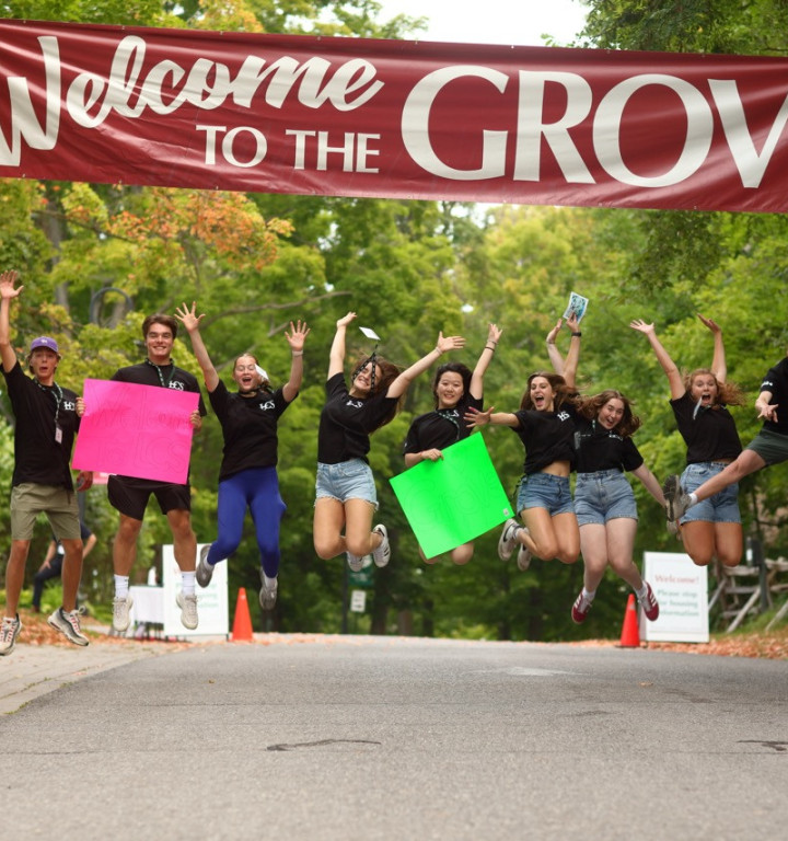 Vor Begeisterung in die Luft springende Schüler unter einem großen Banner auf dem Welcome to the Grove steht